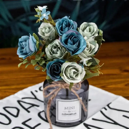 A bouquet of silk fake flowers featuring blue peony and tea rose in a dark purple vase with green leaves and twine