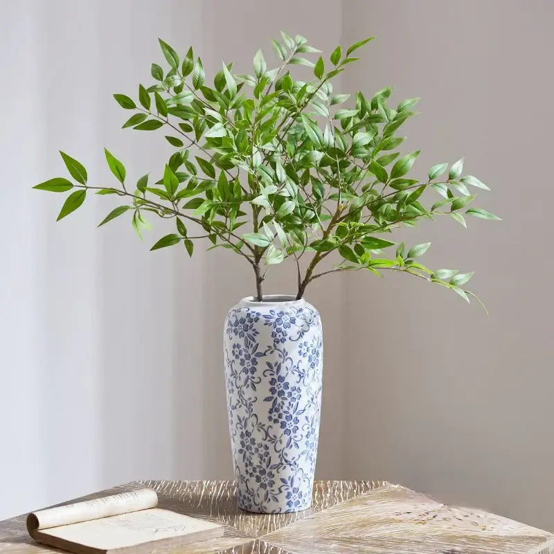 A tall slender white ceramic vase with blue floral pattern holding green foliage for decorbites blue living room decor