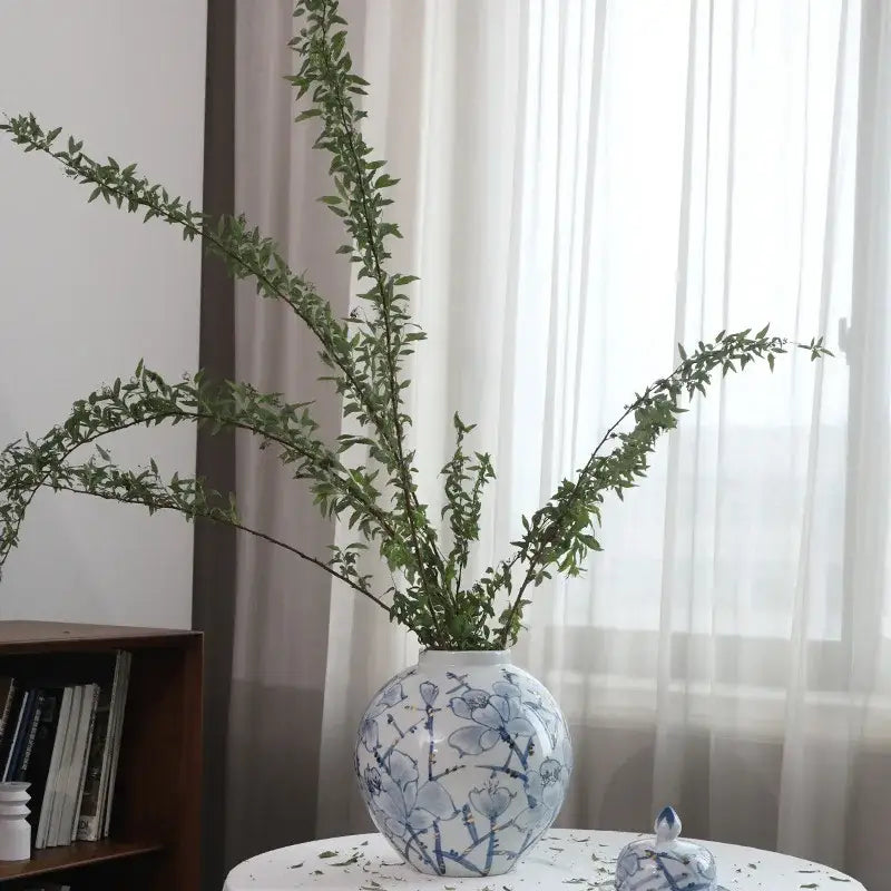 A round blue and white porcelain ceramic vase with floral pattern holding tall green branches for decorbites blue decor