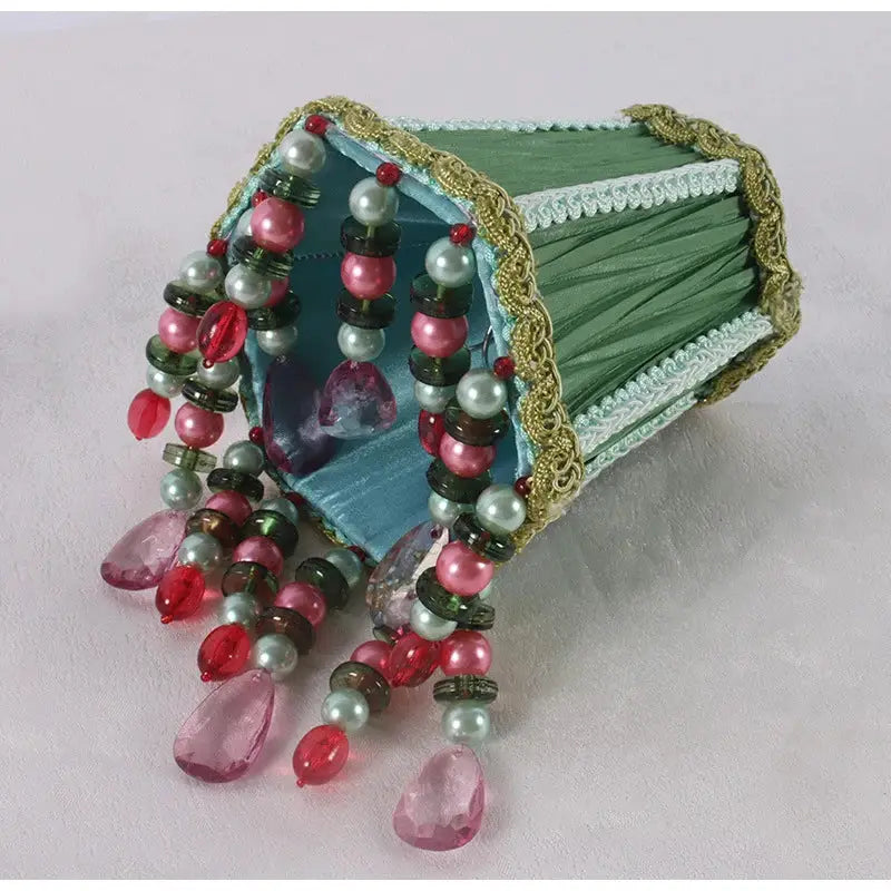 A decorative emerald green cloth jewelry box with gold beading and cascading pink red and pearl-like beads spilling out