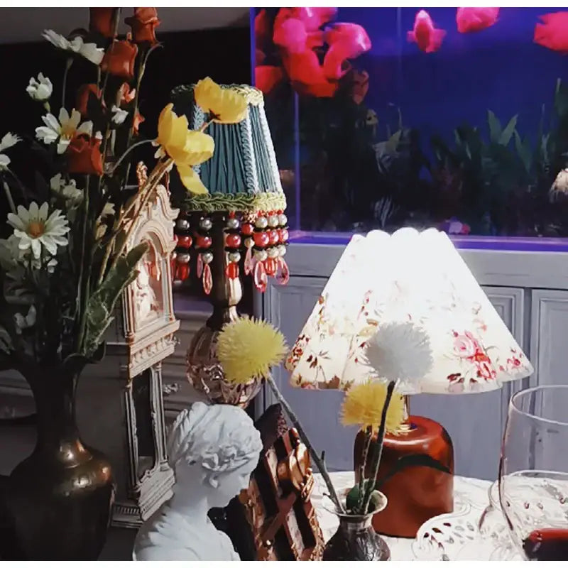 A decorative table lamp with a floral-patterned white shade and an emerald green cloth pleated lampshade with beads