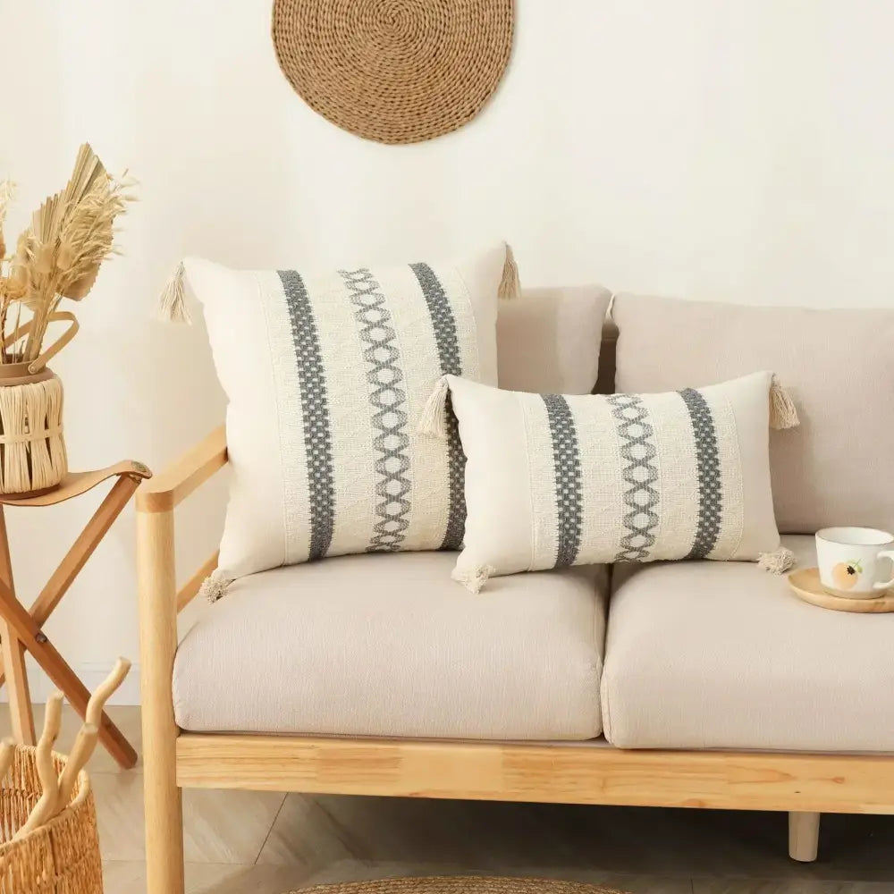 Two cream-colored woven bohemian tassel cushion covers with gray geometric stripes on a light wood sofa frame