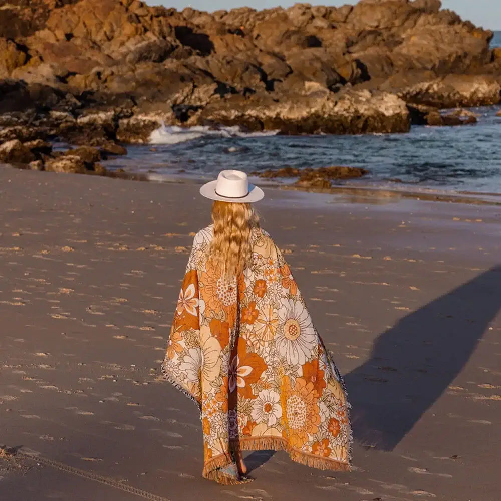A woman walks along a beach wearing a rust and cream bohemian floral leisure blanket with fringe by decorbites