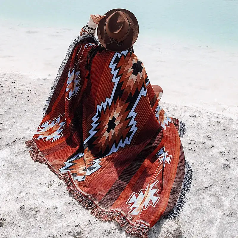 A vibrant orange and brown bohemian knit throw blanket with geometric tribal patterns and fringed edges draped on a beach user