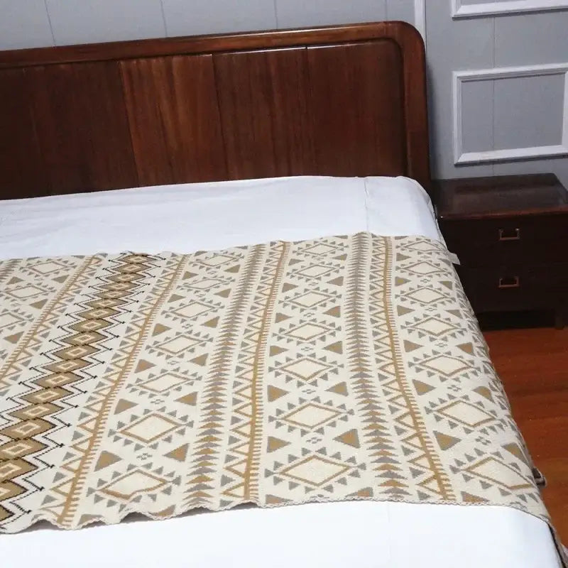 Beige and brown bohemian knitted sofa blanket with geometric pattern and woven texture draped over bed edge decorbites