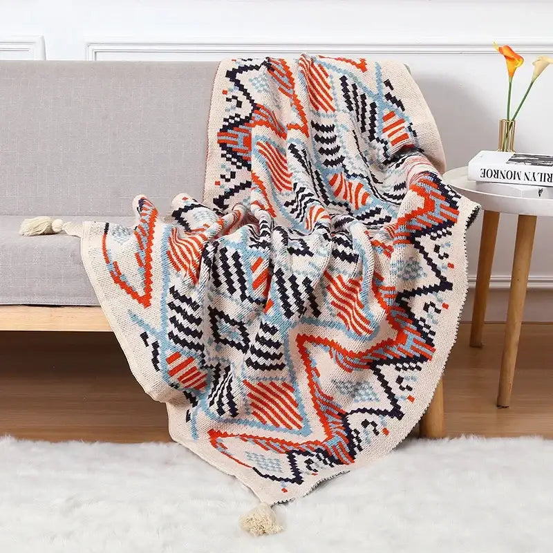 A decorbites bohemian knitted sofa blanket in cream with bold orange black and light blue geometric patterns and tassels