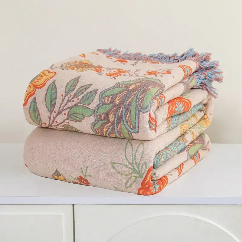 A soft cream-colored decorbites bohemian sofa blanket with vibrant orange and green floral pattern and fringed edges