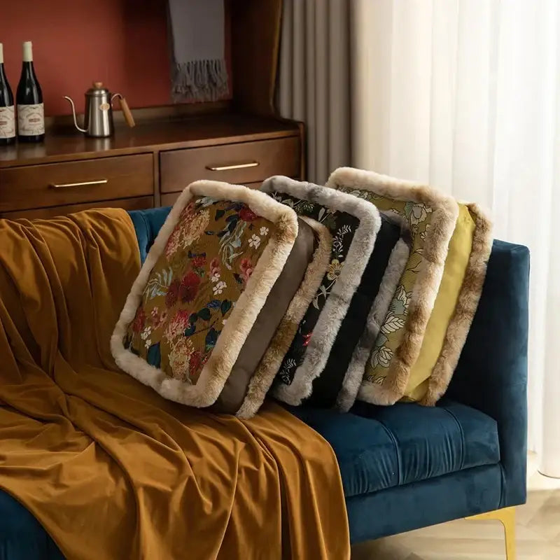 Four rectangular throw pillows with floral patterns and faux fur trim in earthy tones rest on a blue velvet sofa.