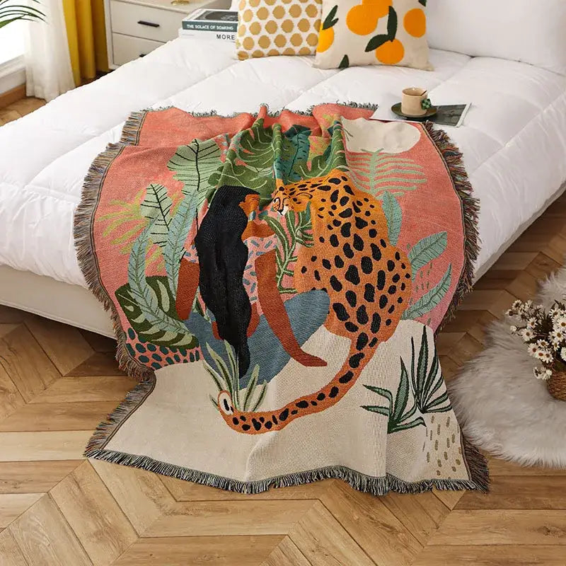 A cozy, woven throw blanket featuring a vibrant jungle scene with an orange and black spotted leopard amidst lush green foliage on a soft peach background.