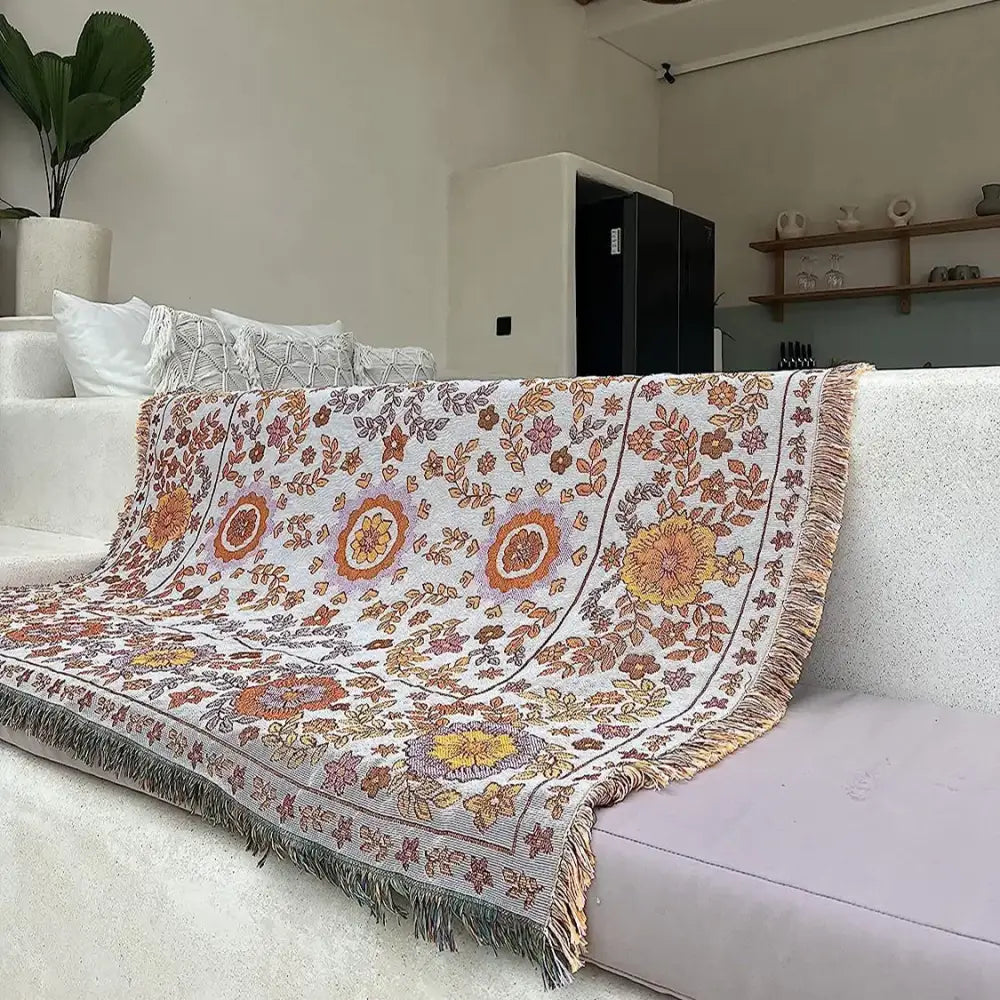 A cozy, off-white woven throw blanket featuring a vibrant floral pattern in shades of orange, yellow, and muted purple, with decorative fringe along its edges.