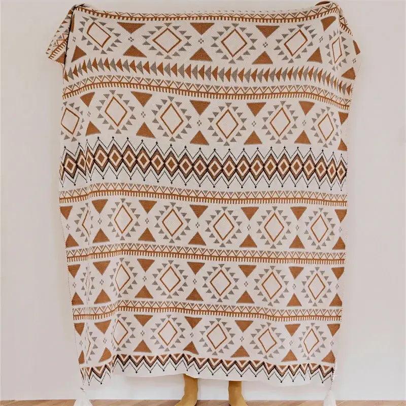 A cozy cream-colored throw blanket featuring a geometric pattern in earthy brown and gray tones, with decorative tassels at the corners.