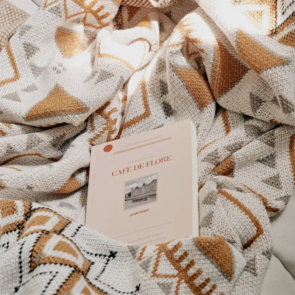A cream and brown boho geometric pattern throw blanket with tassels featuring a soft book on a cozy knitted surface