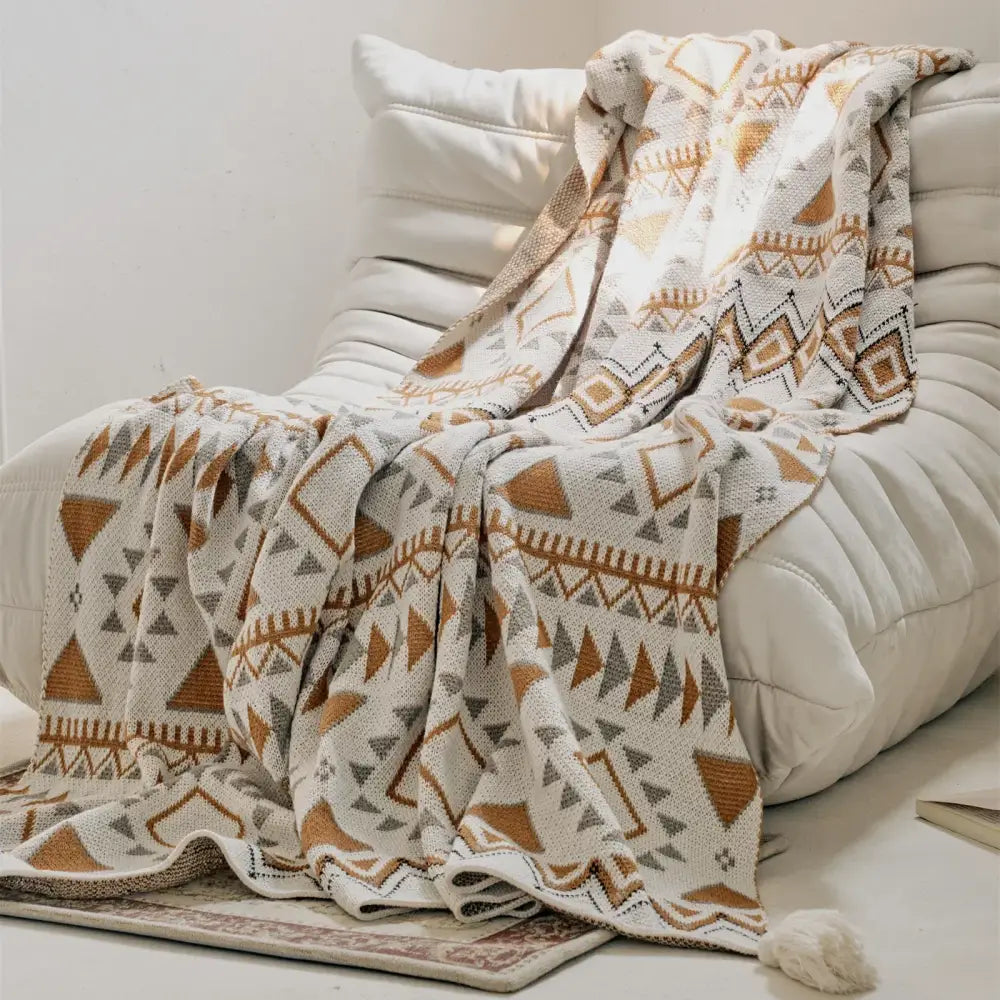 A cozy cream-colored knit throw blanket featuring a geometric pattern in warm terracotta and muted gray tones, with a soft tassel at one corner.