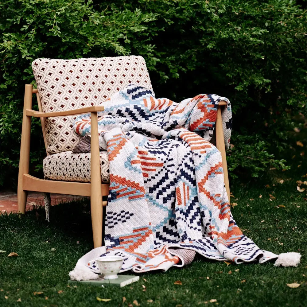 A cozy hand-knitted boho geometric summer blanket in orange navy blue and teal draped over a wooden armchair