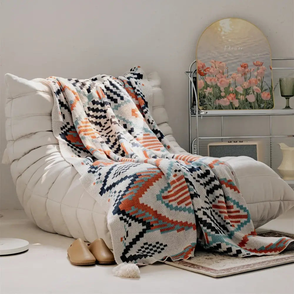 A cozy, knitted throw blanket with a geometric pattern in cream, orange, navy blue, and teal hues draped over a white cushioned chair.