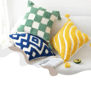 Three colorful, textured throw pillows with tassels, green and white checkered, blue geometric pattern on cream base, and yellow and white striped, all made of plush yarn.