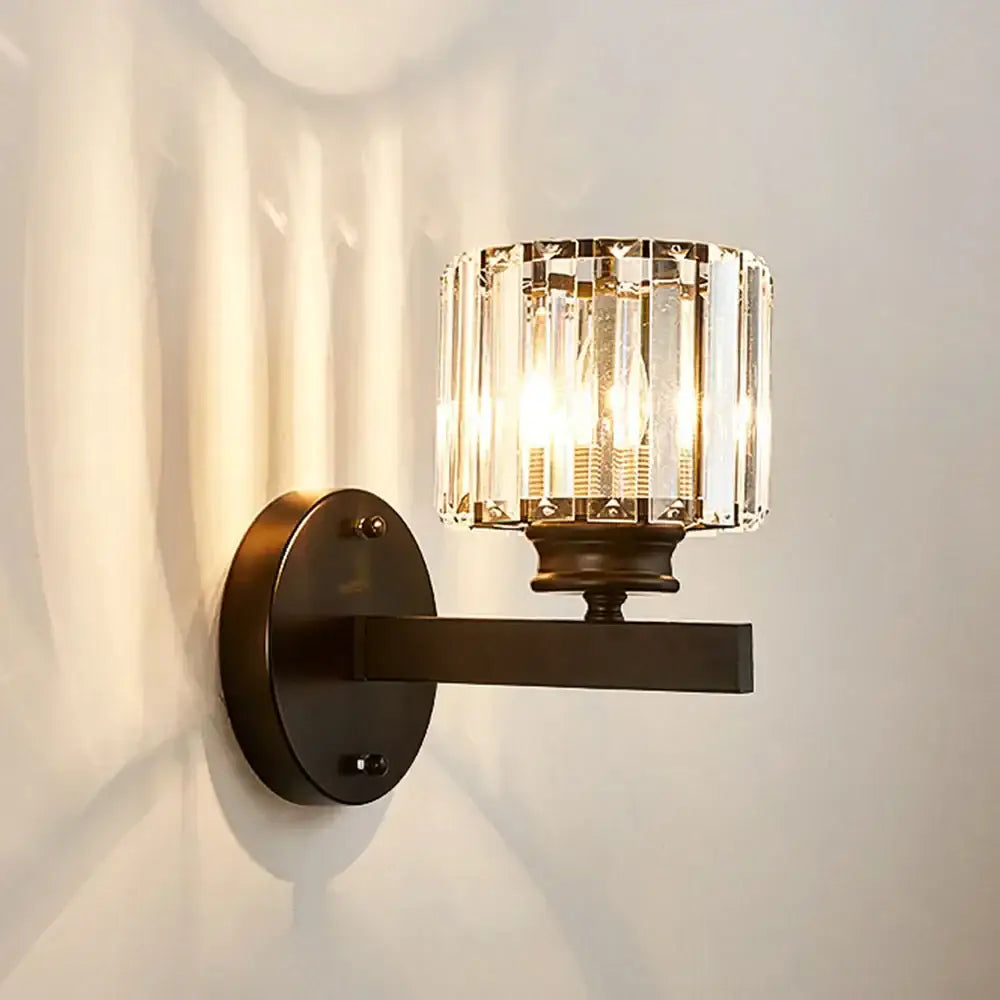 A sleek dark bronze brass decorbites crystal glass wall sconce with a cylindrical faceted crystal glass shade for bedroom lighting