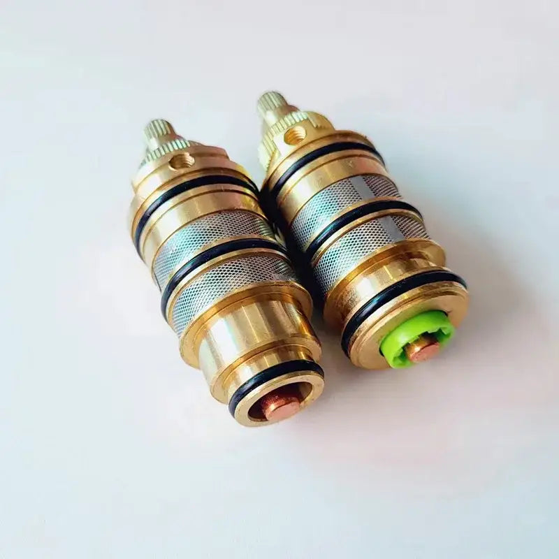 Two brass thermostatic mixer cartridges with meshed bodies, black seals, and one green plastic cap for kitchen use