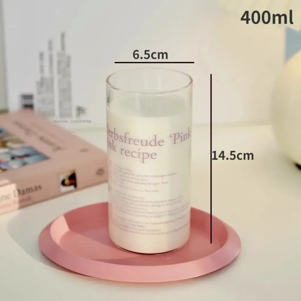 A clear glass coffee mug with white wax candle inside and printed herbsfreude pink ink recipe text on pink tray