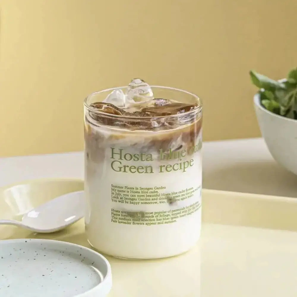 A clear glass coffee mug filled with iced coffee and cream featuring a hosta blue green recipe label and straw