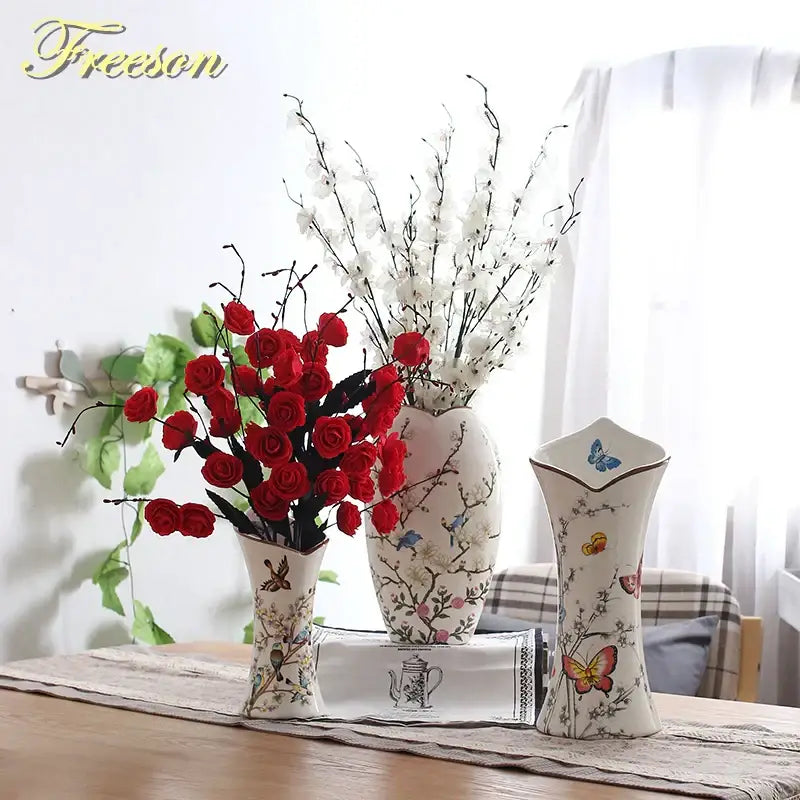 Three decorative bird butterfly porcelain vases with floral and butterfly patterns holding red and white artificial flowers