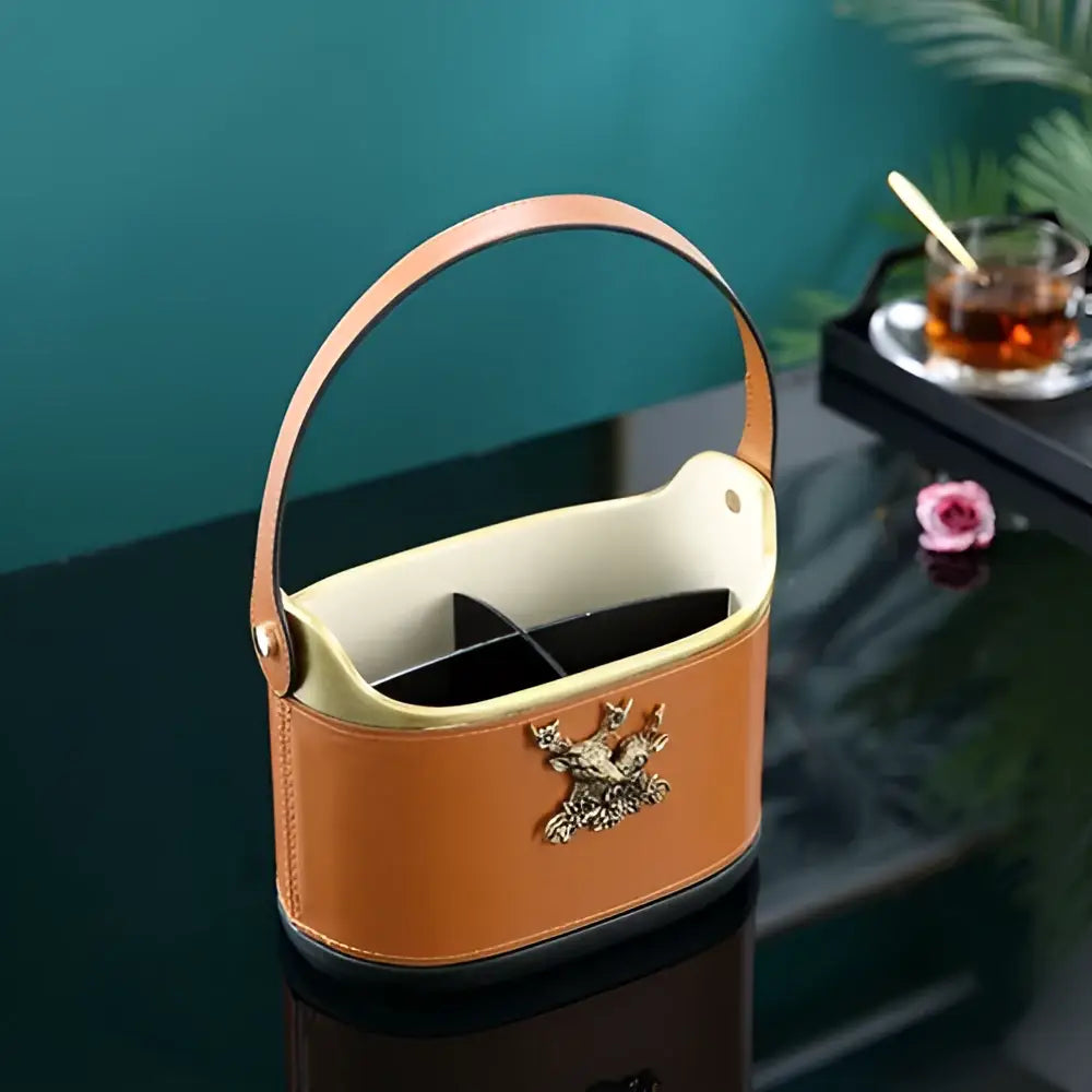 Brown leather desktop storage basket with gold deer emblem, tan leather tea caddy interior, and curved handle for easy carrying