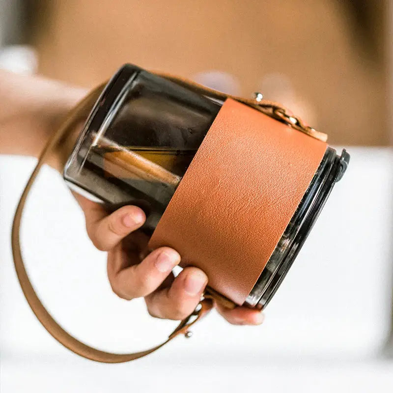 A sleek glass coffee cup with a transparent carafe, tan leather wrap, and matching strap for stylish use