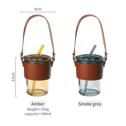 Two insulated glass coffee cups with amber and smoke grey bodies, brown leather sleeves, and gold straws for kitchen use