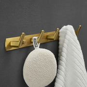 Gold-colored brushed gold robe hooks with three metal hooks holding a white loofah and towel by decorbites