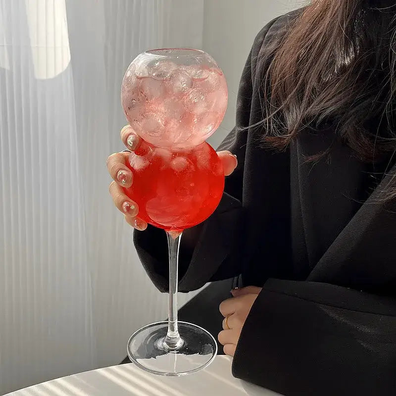 A clear glass bubble ball cocktail glass with a long stem filled with pink liquid and ice from decorbites bubble ball set
