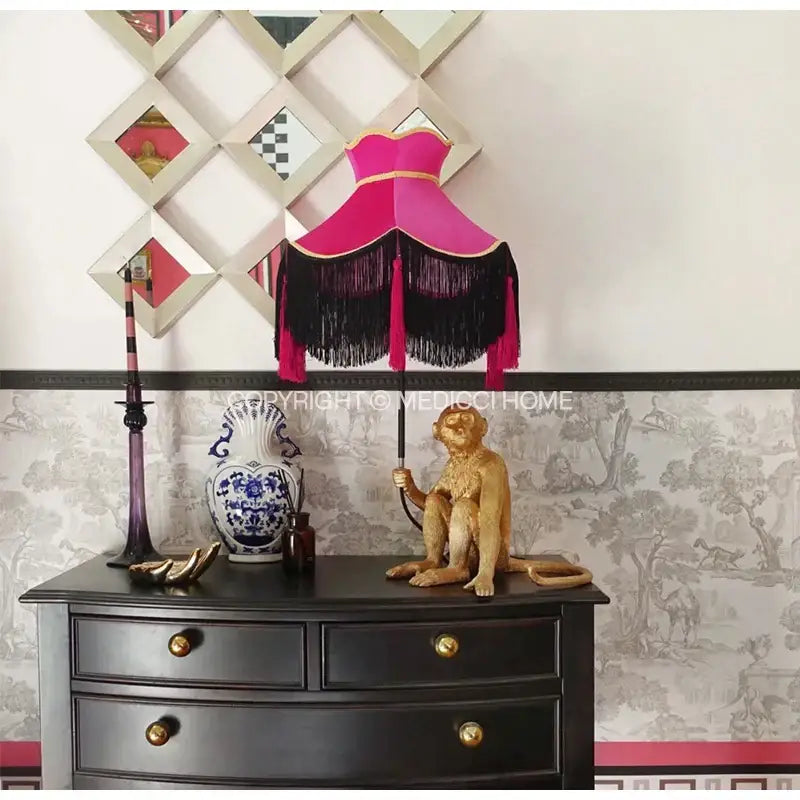 A burgundy wine red velvet scalloped lampshade with black fringe and gold trim atop a golden monkey figurine