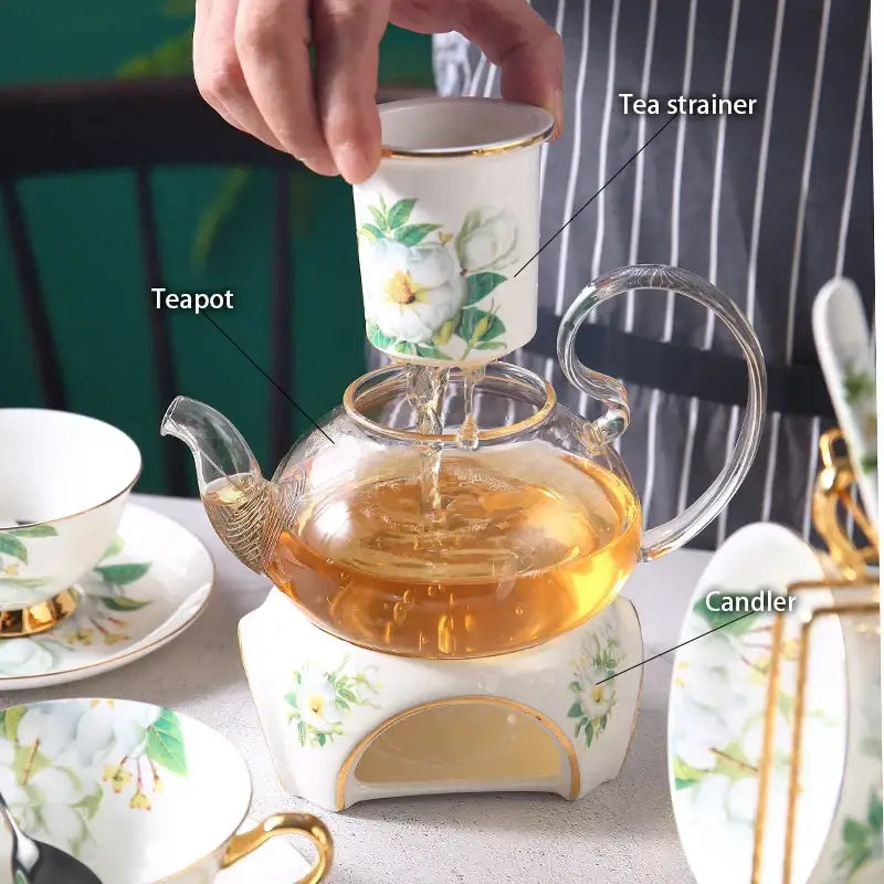 A clear glass teapot with gold trim filled with amber tea on a white ceramic stand in the decorbites camellia floral tea set