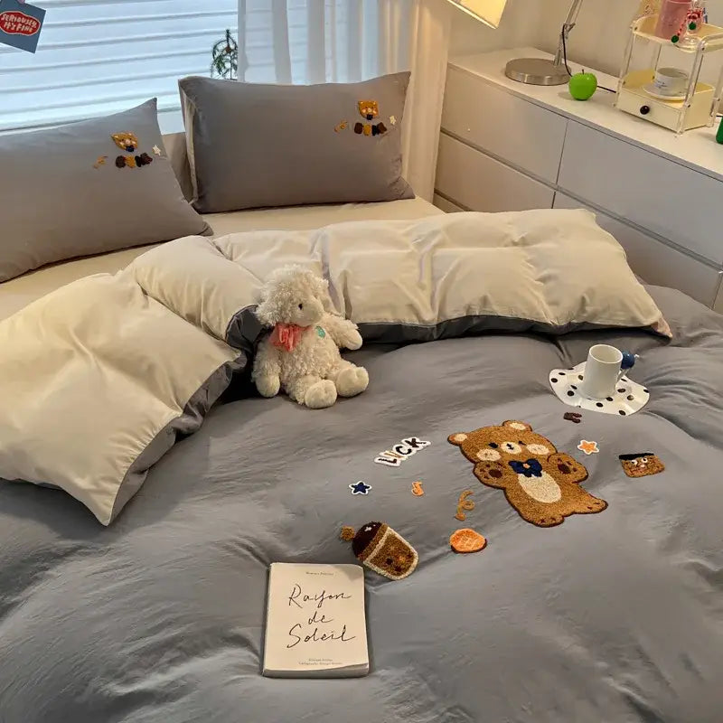 Gray plush teddy bear with pink scarf sitting on bed with cartoon pattern embroidered cream bedding sets decorbites decor