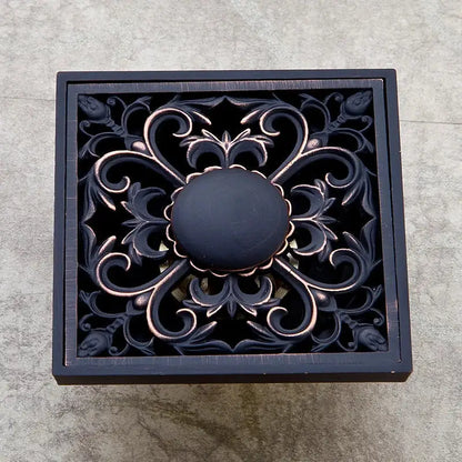 Square bronze-finished decorbites black floor drain with intricate floral scrollwork and central matte black knob for bathroom use