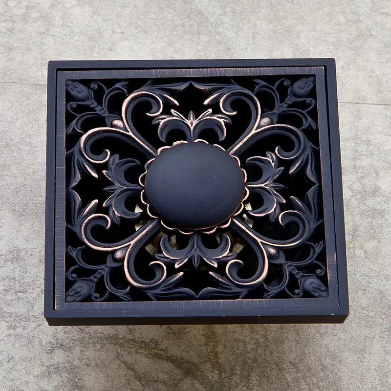 Square bronze-finished decorbites black floor drain with intricate floral scrollwork and central matte black knob for bathroom use