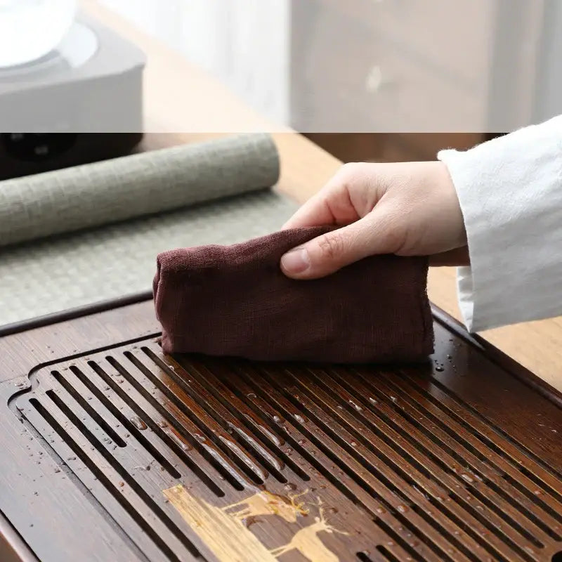 A hand holding a dark brown soft-textured cloth used with a carved deer bamboo tea tray drainage board