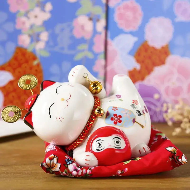 White ceramic maneki neko piggy bank with gold coins and bells on red floral cushion by decorbites ceramic maneki
