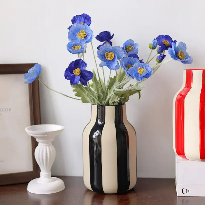 A sleek black and cream striped ceramic vase holding vibrant blue artificial poppies with yellow centers