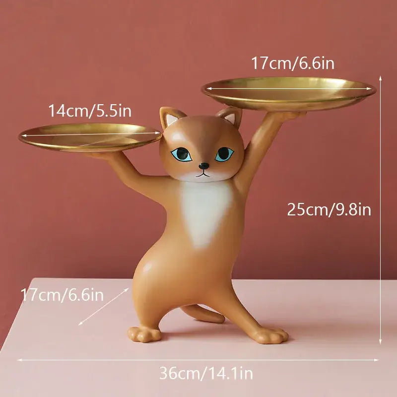 A tan resin cat figurine with blue eyes balancing golden plates in a ceramic white and beige cat figurines storage tray