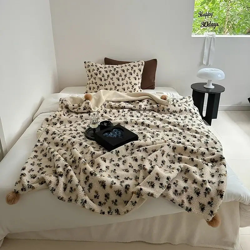 Cream-colored sheep rollie loop fleece blanket with black floral pattern and fluffy pom-pom tassels for bedroom use