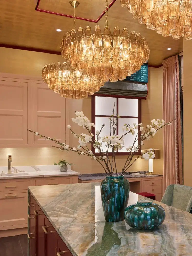 A tall glossy teal ceramic vase with textured surface on marble countertop complements decorbites large pink glass chandelier lighting