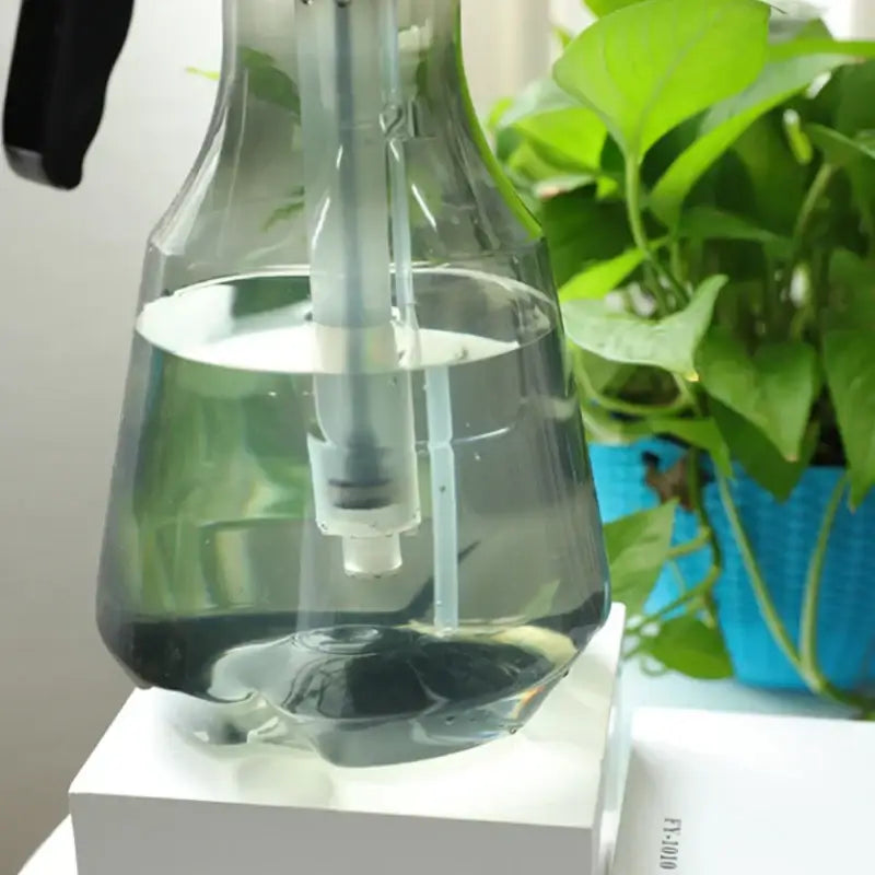 Clear glass watering sprayer with black hand trigger and white internal parts filled halfway for garden planting use