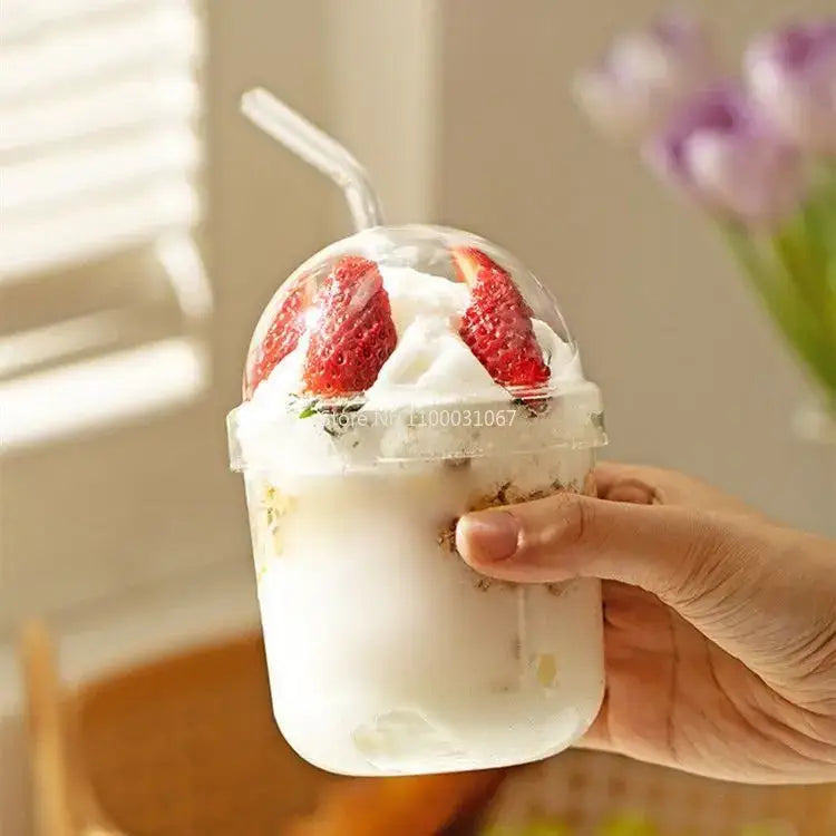 Clear glass bobo cup with ribbed lid and curved straw containing white milkshake garnished with two red strawberries