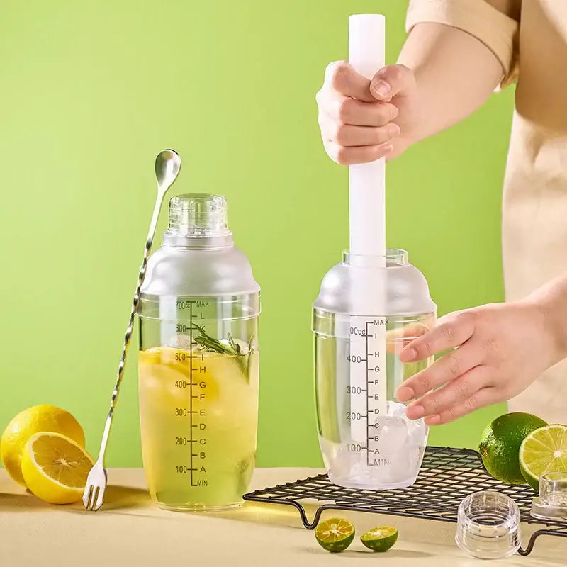 Clear glass cocktail shaker with silver lid and measurement markings beside a silver-toned bar spoon for beverage mixing