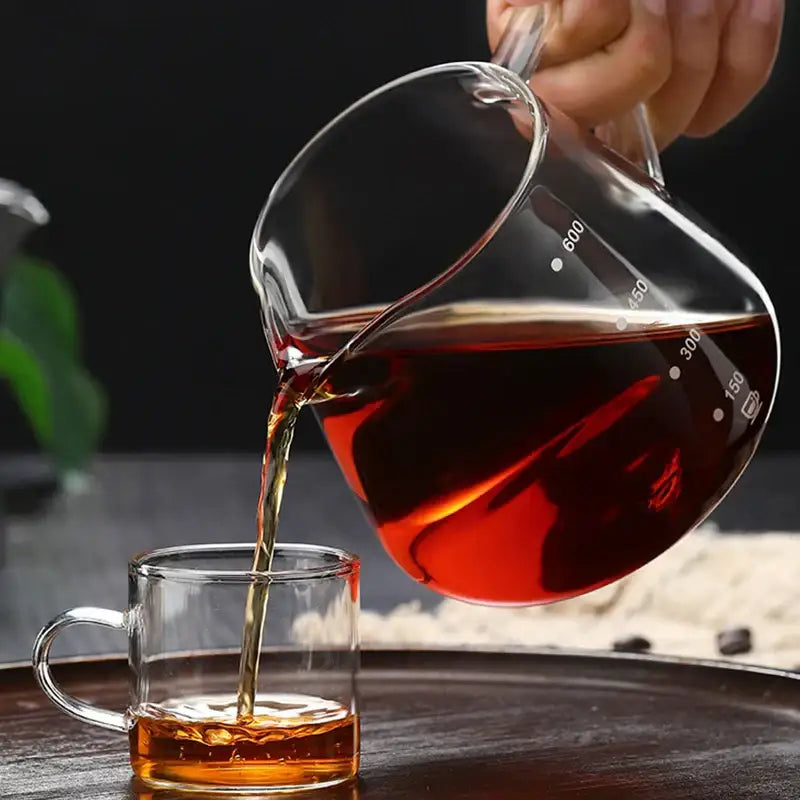 Clear glass coffee pot with measurement markings pouring dark amber tea into a matching glass coffee mug