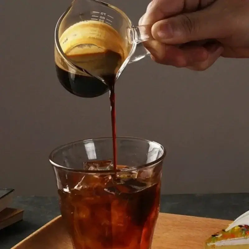 Clear glass espresso measuring cup with handle and measurement markings pouring dark coffee over ice in a glass
