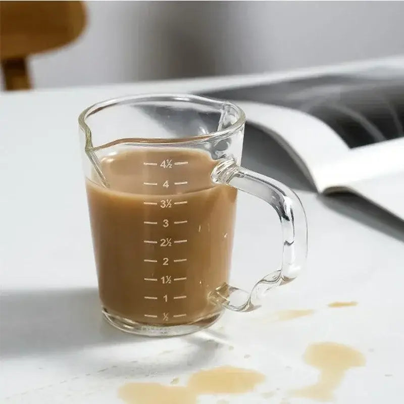 Clear glass espresso measuring cup with handle filled with brown liquid and marked with measurement lines for precise use