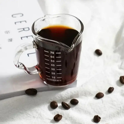 Clear glass espresso measuring cup with handle and white milliliter markings filled with dark coffee