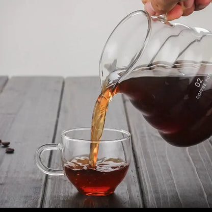 Clear glass cloud glass coffee kettle with silver handle pouring dark brew into matching clear glass mug with measurement markings