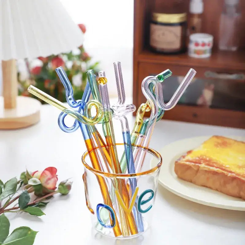 A clear glass cup with a gold rim holds colorful transparent bendy straws with a flower handle stir bar